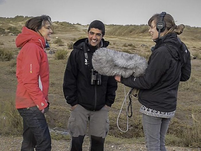 Ajay Tegala recording for Living World Trai in a red coat and Ajay in a black coat, are with a sound recordist holding a big, fluffy microphone, they are on a shingly beach with sand dunes behind them and a few seals just visible dotted around in the background, the sky is grey