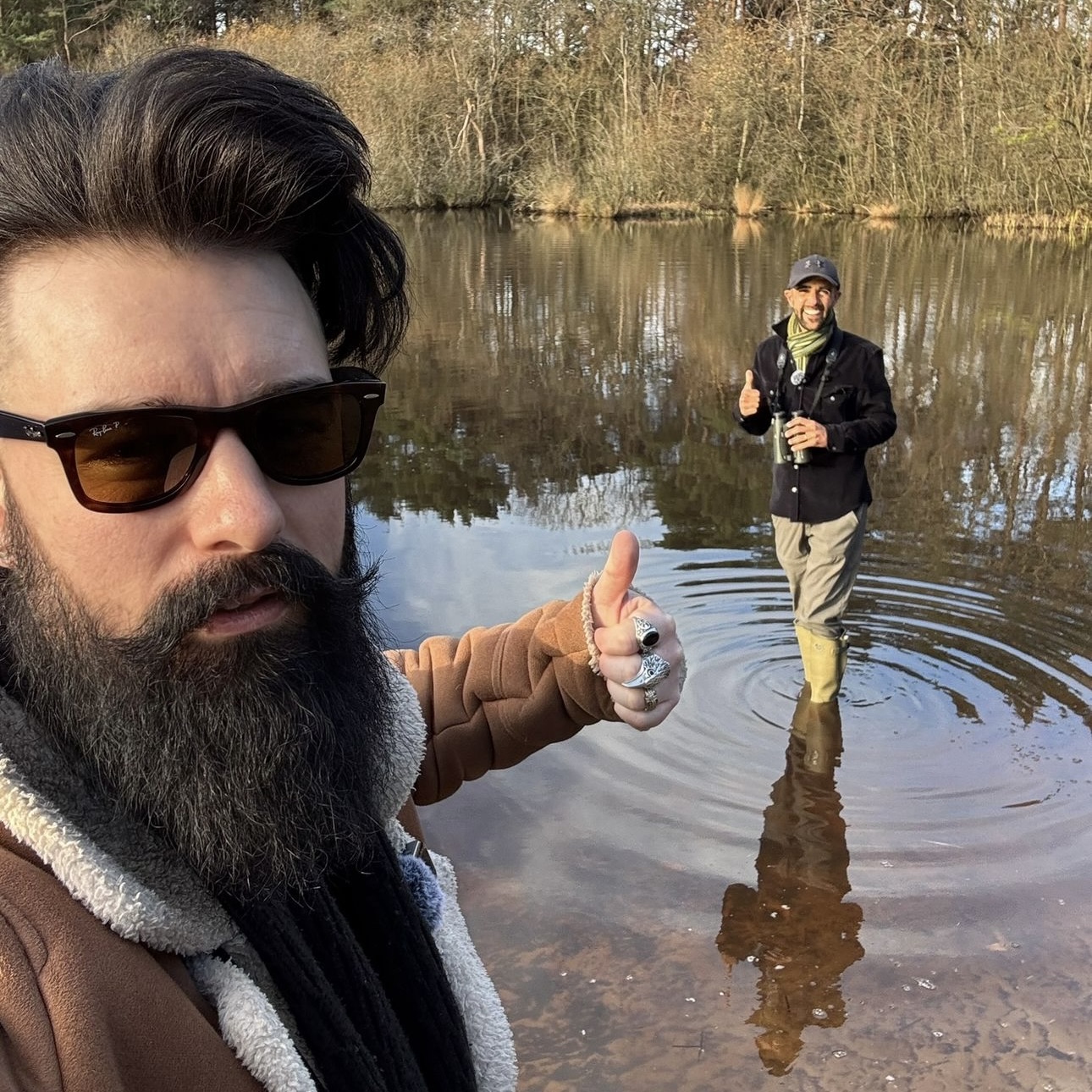 Matt with sunglasses, long black beard and styled hair stands in foreground with thumb up, in the background is a lake with trees beyond, Ajay is stood in the water, wearing wellies, with his thumb up, wearing a baseball cap