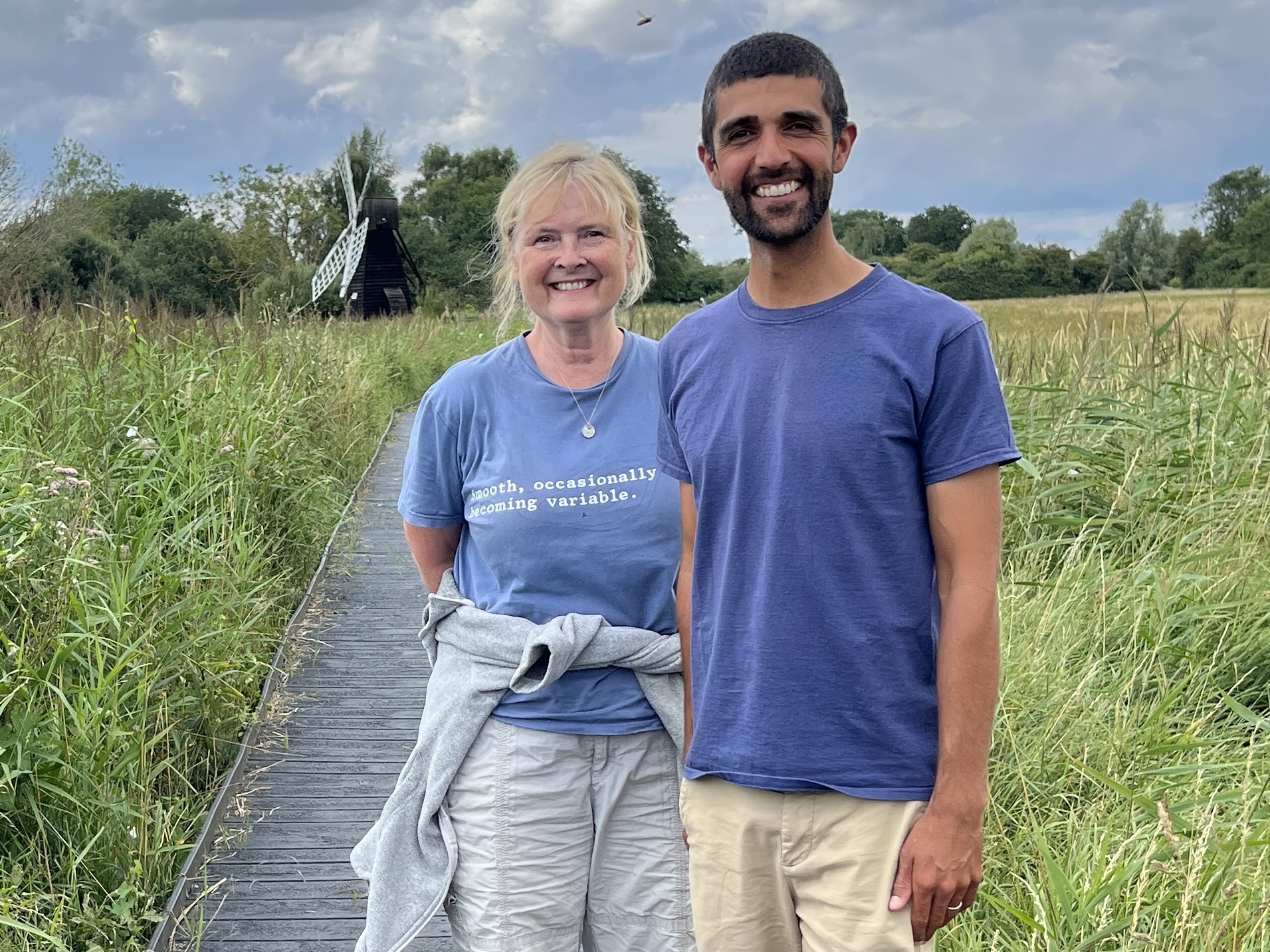 2025-07-21 Martha Kearney and Ajay Tegala (2025) Martha and Ajay stand side by side, both wearing blue T-shirt, pale-coloured shorts and smiling, behind them is lush green vegetation, blue sky with grey-white, fluffy clouds and a windmill in the distance, with a boardwalk leading towards it, there is a line of trees on the horizon