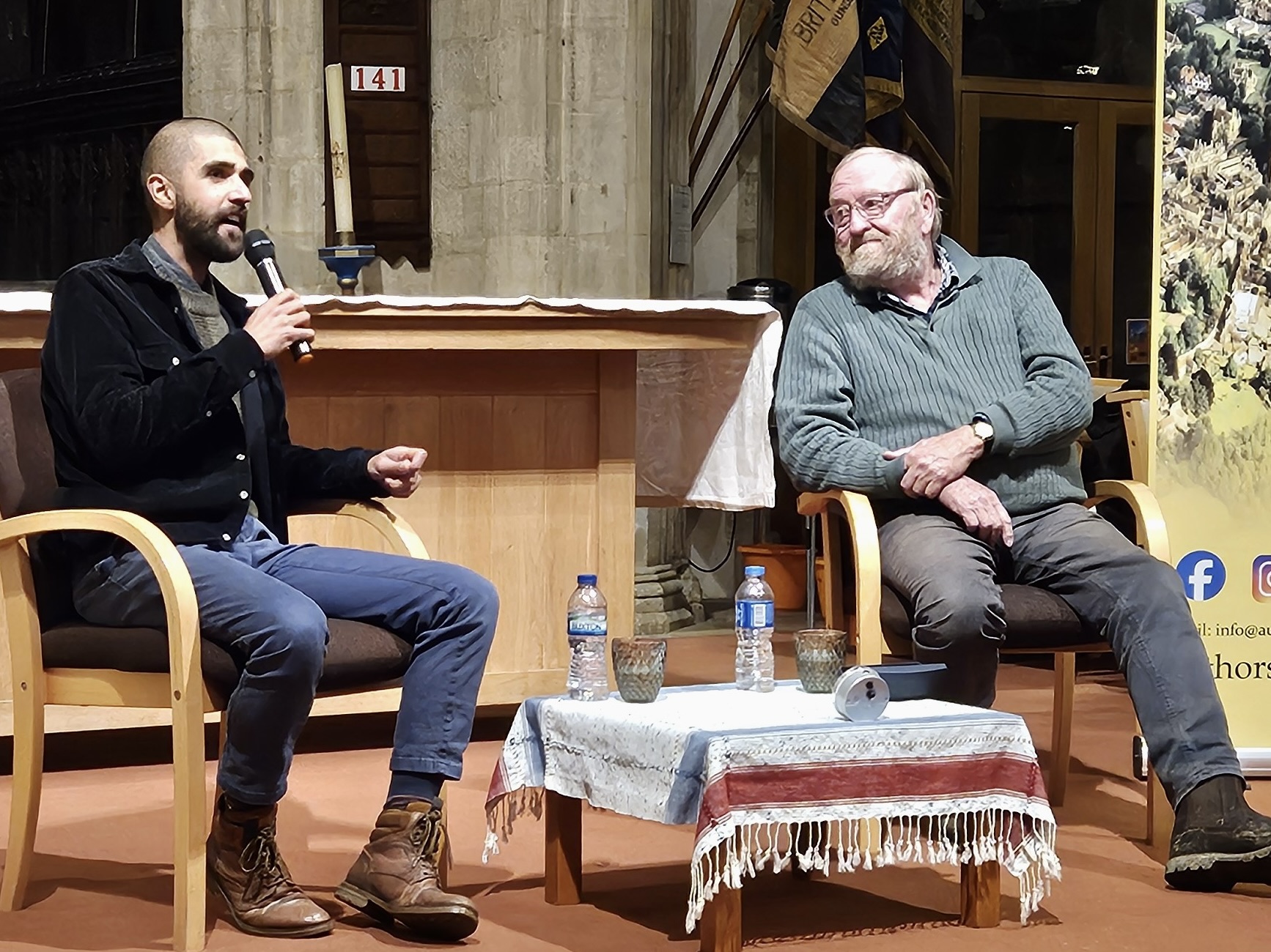 Ajay Tegala and Francis Pryor (Authors in Oundle) Two men sat on chairs in a church, Ajay is on the left talking into a microphone, Francis is on the right looking towards him, in front of them is a low table with bottles of water and a clock on top of a table cloth.