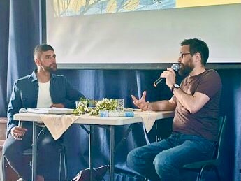 Ajay Tegala interviews Michael J Warren at Deepings Literary Festival Two men sat by a table, one holding a microphone