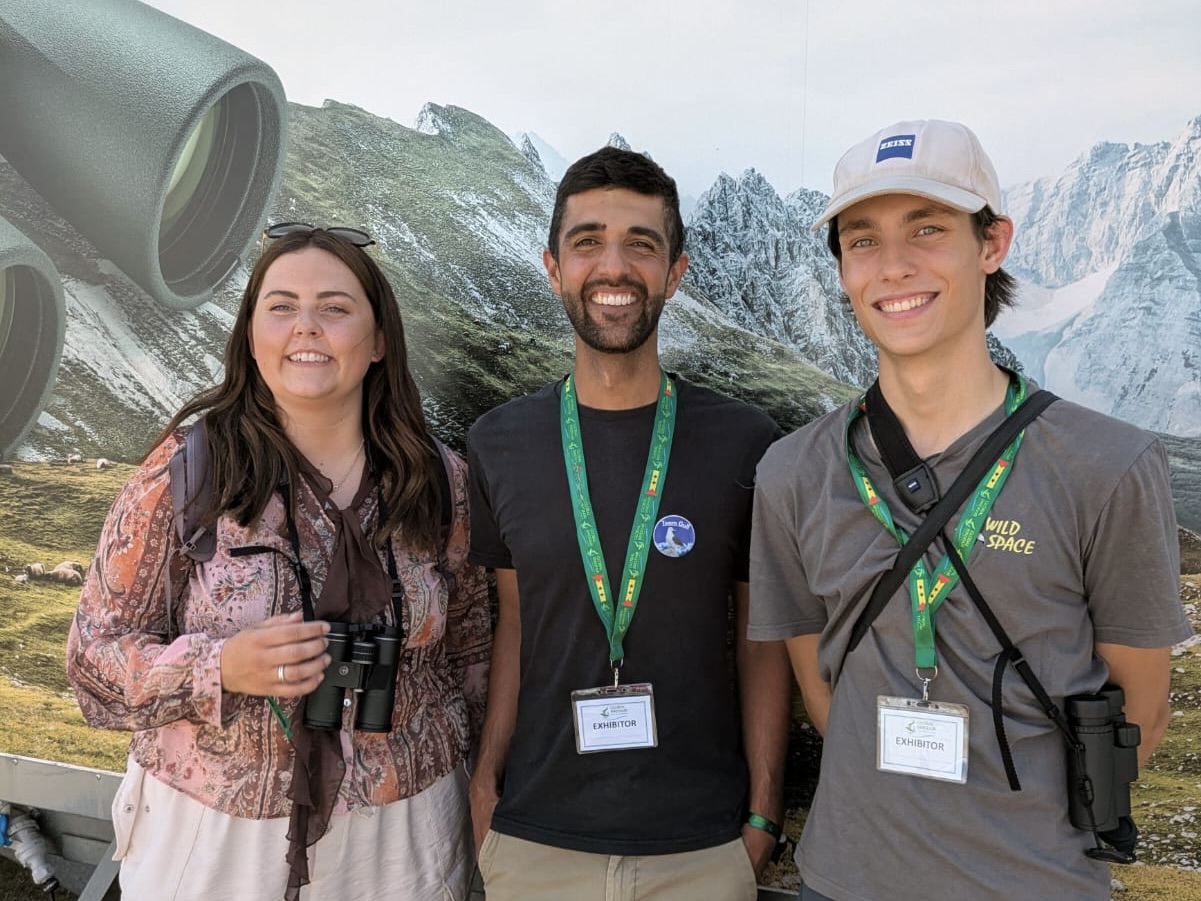 Ajay Tegala with Emily Askew and Ben Rumsby at BirdFair Three smiling young people in front of a poster of mountains