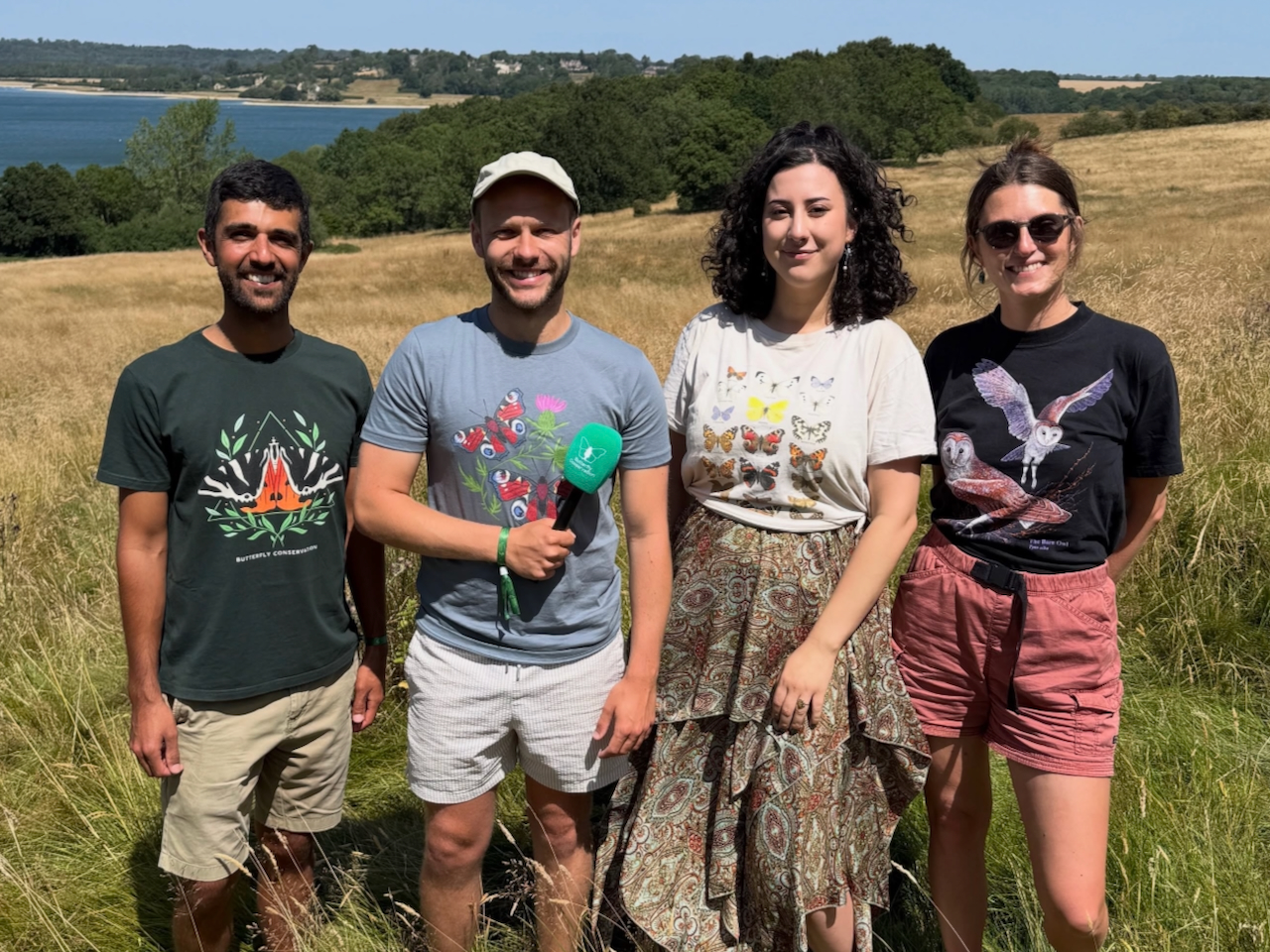 Ajay Tegala presents alongside other Butterfly Conservation ambassadors Four smiling young people outside with grass, trees, water and blue sky behind them, there are two men, one holding a microphone and two ladies, all are wearing summer clothes and smiling