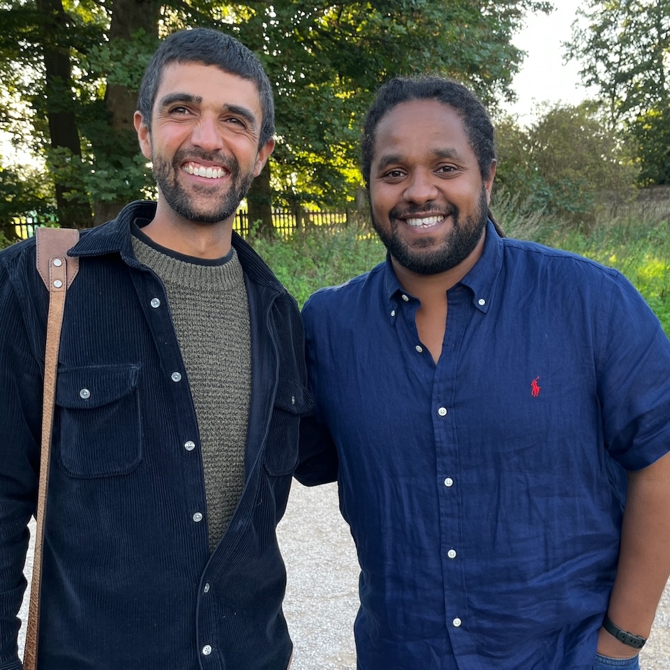 Two men stood together smiling outdoors in front of some trees with the sun's low rays shining through