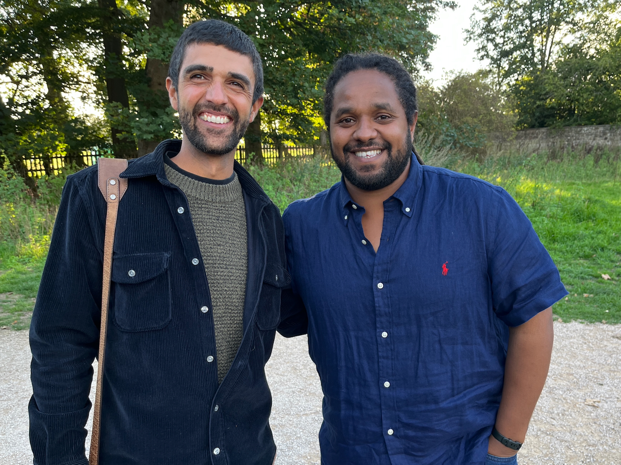 Ajay Tegala and Hamza Yassin Two men stood together smiling outdoors in front of some trees with the sun's low rays shining through